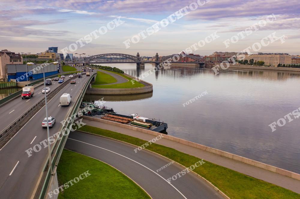 На фото изображен Большеохтинский мост в Санкт-Петербурге, соединяющий исторический центр с промышленными районами. На переднем плане – современные магистрали, вписанные в городскую архитектуру. Река Нева отражает спокойное небо, создавая умиротворяющую атмосферу. Мост и окружающие улицы символизируют сочетание истории и современности города.